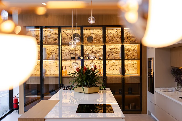 Adega com portas de vidro e parede de pedra iluminada, ilha com cooktop e pendentes