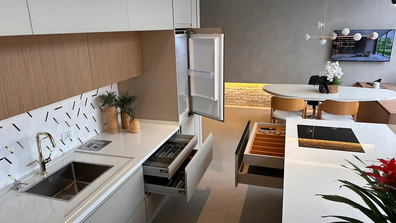 Cozinha do showroom com cuba sob o tampo, torneira dourada, lixeira embutida na bancada, gavetas abertas com organizadores, geladeira embutida e cooktop na ilha